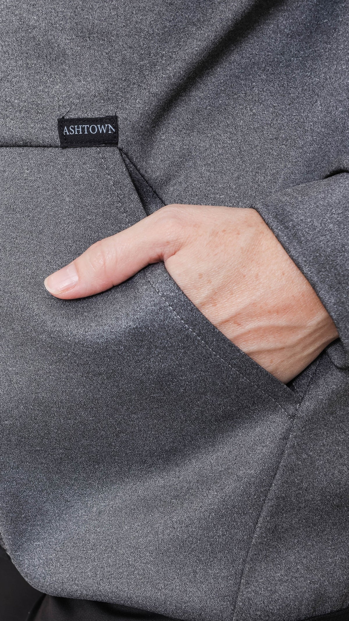 Close-up of a grey Ashtown Clothing performance hoodie showing a hand in the front pocket with a branded label.