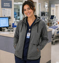 A smiling nurse wearing an Ashtown Clothing grey moisture-wicking full-zip hooded jacket over navy blue scrubs.