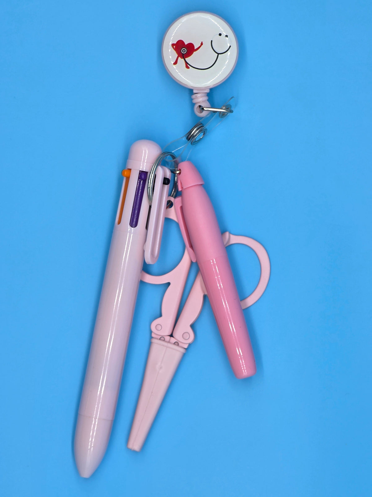 Ashtown Clothing pink retractable badge reel, multi-color pen, and medical scissors set on a blue background.